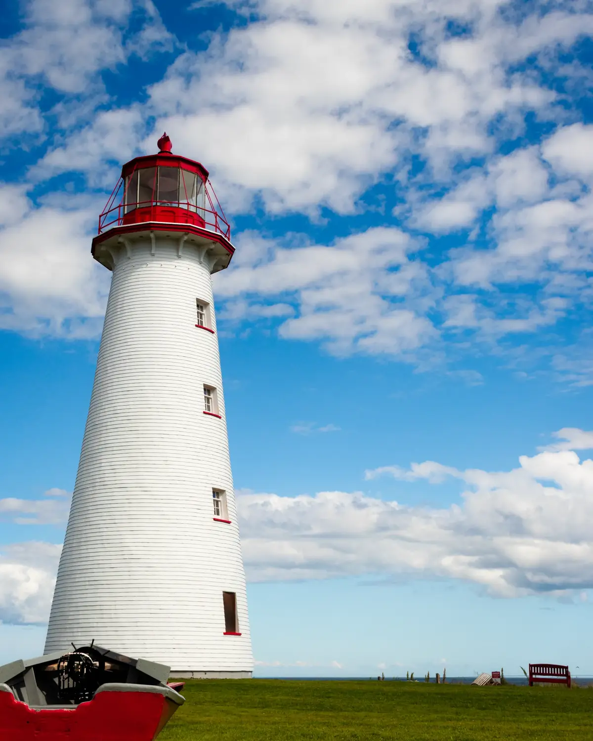 The white, shingled Point Prim Lighthouse under a blue sky full of cloud, with an old red boat on the grass beside it.
