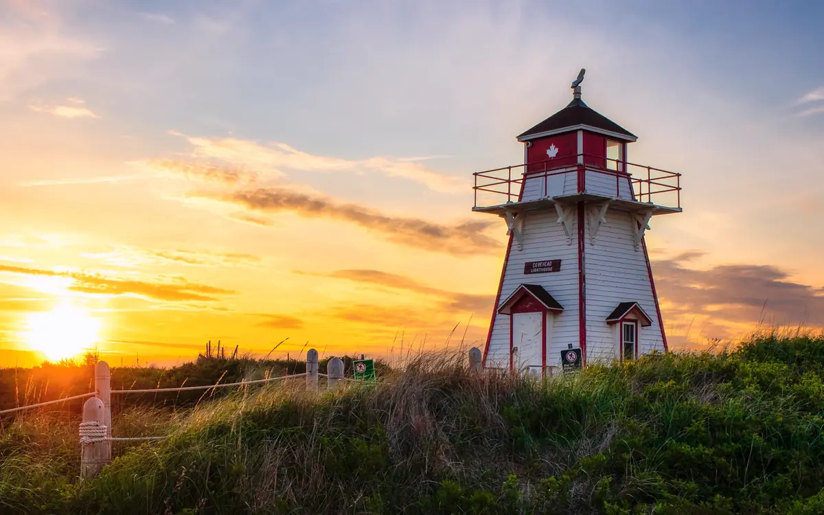 The red and white Covehead Harbour Lighthouse on a grassy dune at sunset, with the sun low beside it.